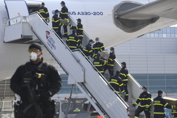 A police officer holds a weapon as firefighters unload an airplane after its arrival at the Vaclav Havel Airport in Prague, Friday, March 20, 2020. The airplane brought medical aid and protective materials against coronavirus from China. (Michal Kamaryt/CTK via AP)