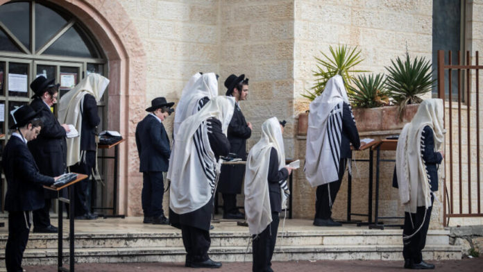 Jewish men pray outside a synagogue in the city of Beitar Illit in Judea and Samaria, on March 29, 2020 (Photo by Aharon Krohn/Flash90)