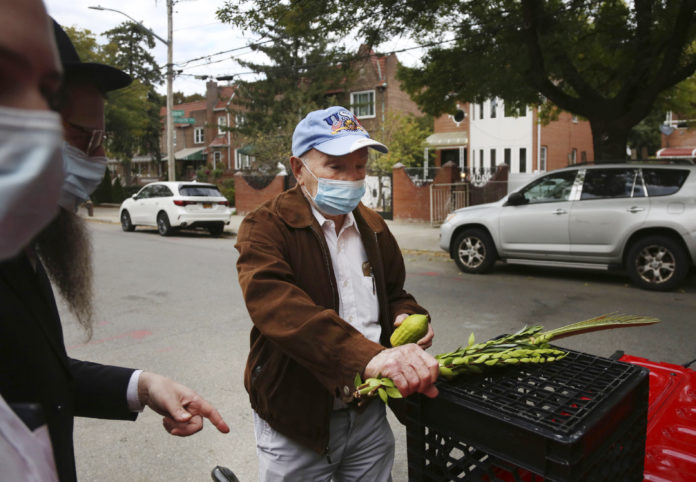 Holocaust survivor Leon Sherman holds a lulav, a collection of palm, myrtle and willow branches, and an etrog, a citrus fruit, as he recites the blessings in front of his home in the Queens borough of New York, Monday, Oct. 5, 2020. In an effort to bring the Jewish fall harvest traditions safely to those isolated by the coronavirus, Rabbi Eli Blokh, of the Chabad of Rego Park Jewish and Russian Community Center, built a mobile sukkah, a temporary shelter where Jews gather to celebrate Sukkot, in the back of a red pickup, making several house calls each day during the weeklong festival to people like Sherman. (AP Photo/Jessie Wardarski)