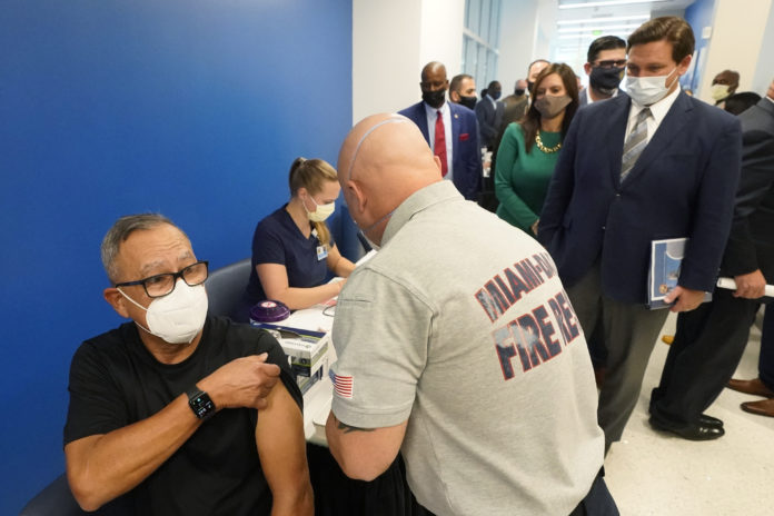 FILE - In this Jan. 4, 2021, file photo, Florida Gov. Ron DeSantis, right rear, watches as Carlos Dennis, left, 65, rolls up his sleeve so that Miami-Dade County Fire Rescue paramedic, Capt. Javier Crespo, can administer a COVID-19 vaccine shot at Jackson Memorial Hospital in Miami. Florida was one of the first states to throw open vaccine eligibility to members of the general public over 65, leading to rumors that tourists and day-trippers are swooping into the state solely for the jab. (AP Photo/Wilfredo Lee, File)