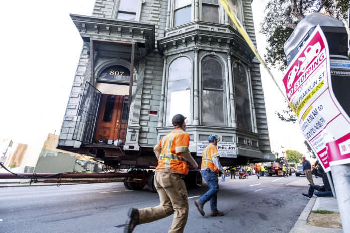 Workers pass a Victorian home as a truck pulls it through San Francisco on Sunday, Feb. 21, 2021. The house, built in 1882, was moved to a new location about six blocks away to make room for a condominium development. According to the consultant overseeing the project, the move cost approximately $200,000 and involved removing street lights, parking meters, and utility lines. (AP Photo/Noah Berger)