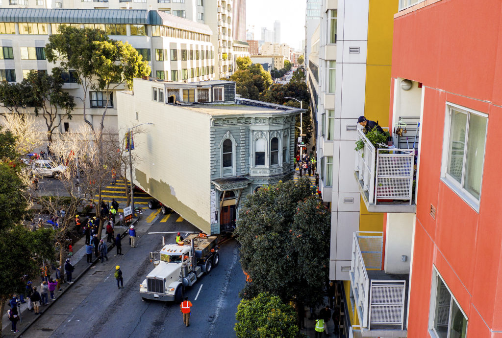 A man watches from a balcony as a truck pulls a Victorian home through San Francisco on Sunday, Feb. 21, 2021. The house, built in 1882, was moved to a new location about six blocks away to make room for a condominium development. According to the consultant overseeing the project, the move cost approximately $200,000 and involved removing street lights, parking meters, and utility lines. (AP Photo/Noah Berger)